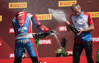 A racer and team member spray champagne on a podium in ZXMOTO team colours.