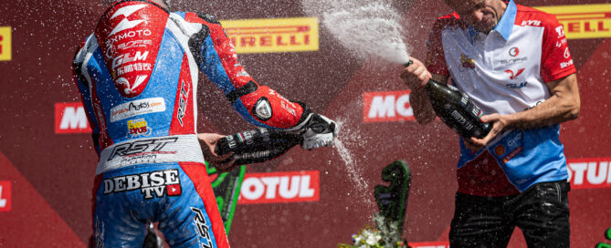 A racer and team member spray champagne on a podium in ZXMOTO team colours.