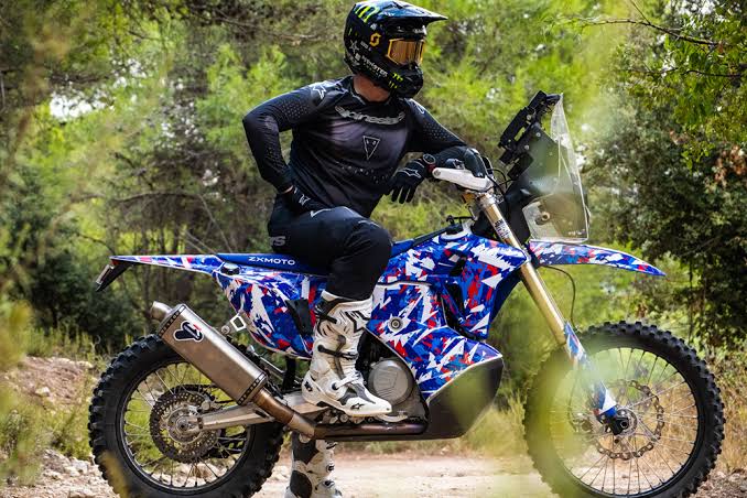 A helmeted rider sits on a blue, red and white ZXMOTO rally motorcycle on a wooded trail.