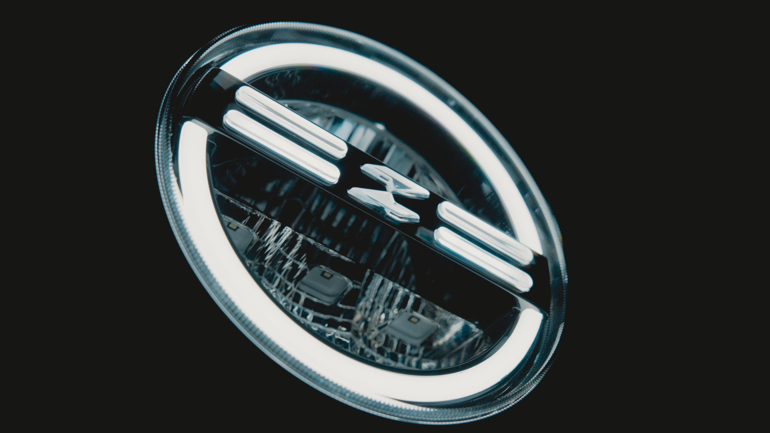 Close-up of a round motorcycle headlight with a bright outer light ring, horizontal light bars and a central emblem.