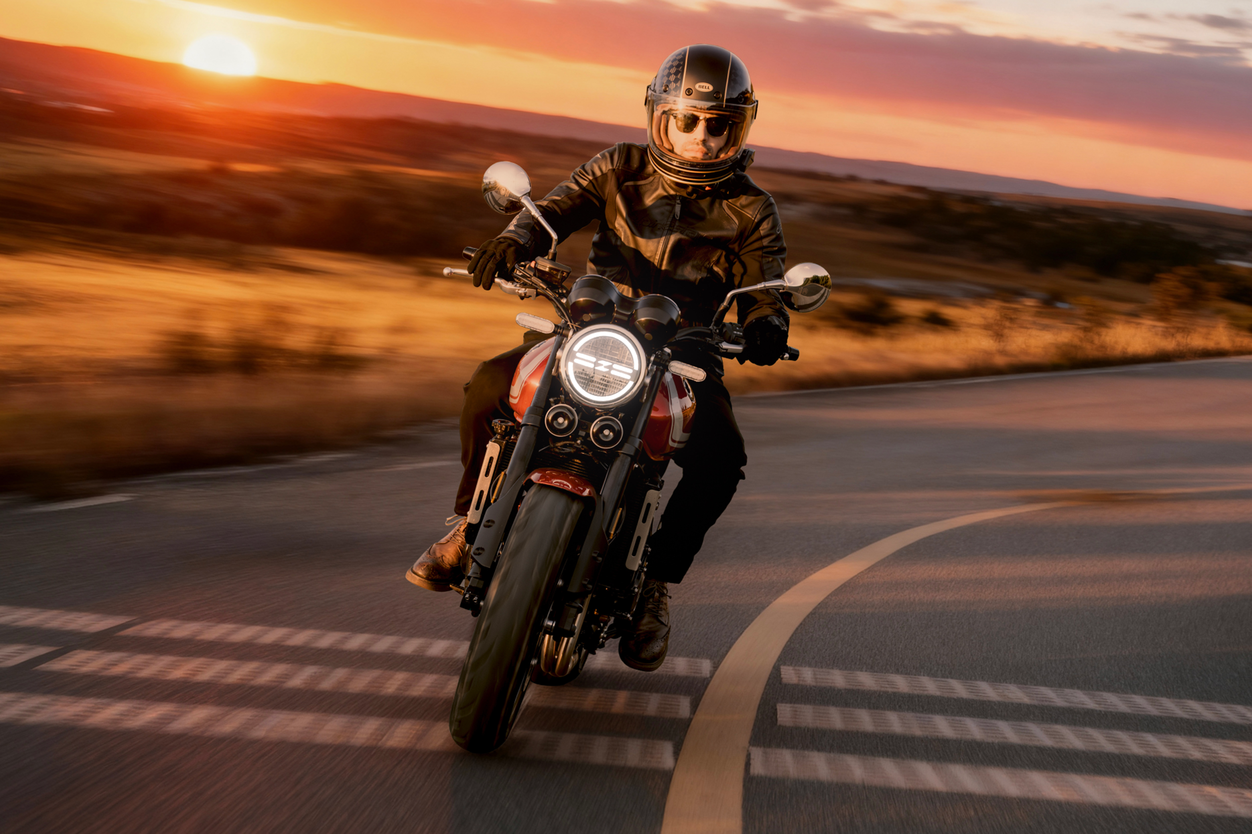 A helmeted rider leans a red 500F through a paved bend with its round headlight lit against an orange sunset.