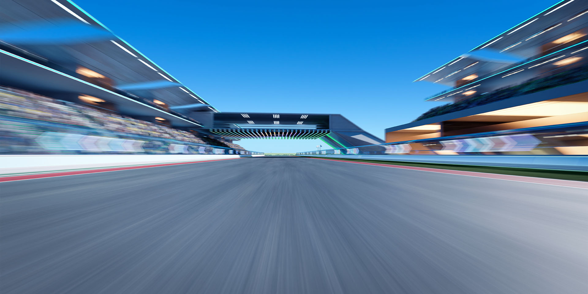 An empty race circuit straight with blurred grandstands and pit buildings beneath a bright blue sky.