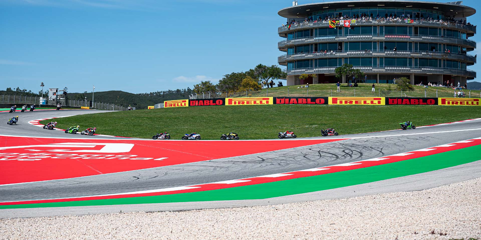 A pack of motorcycle racers rounds a circuit bend beside red and green run-off areas and a large trackside building.