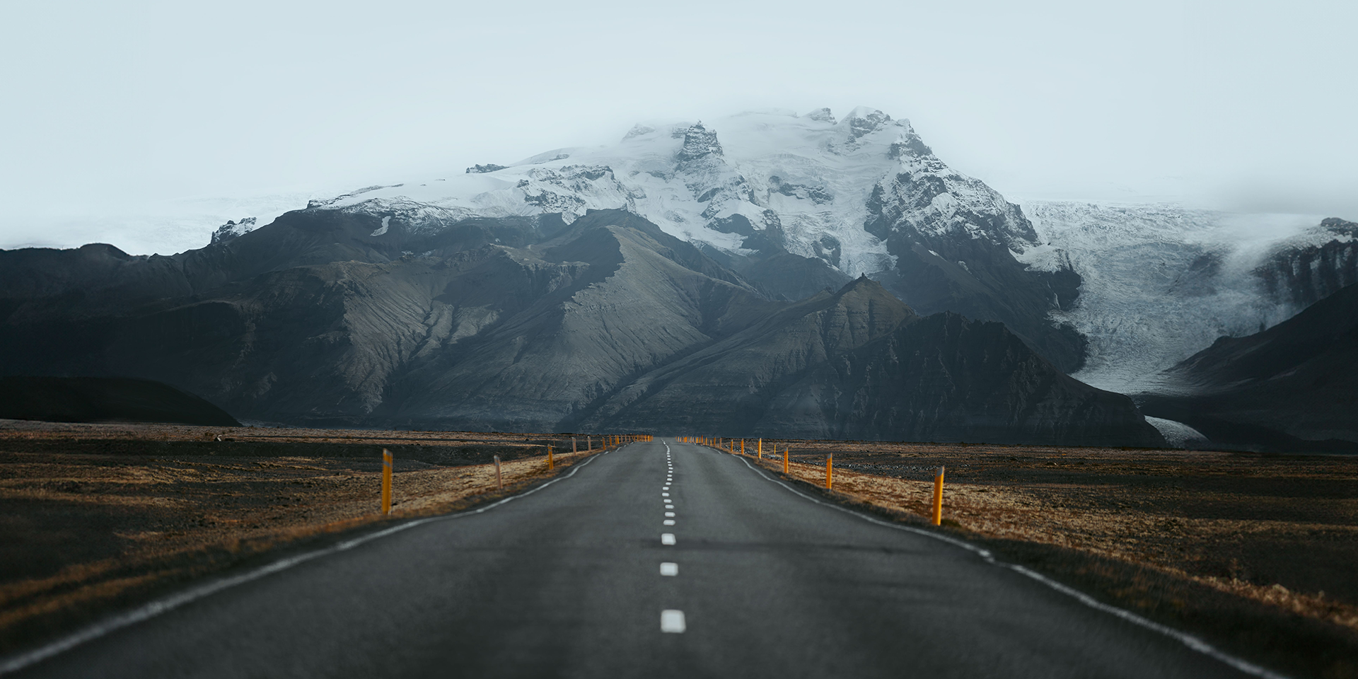 A straight paved road leads towards snow-covered mountains beneath low cloud.