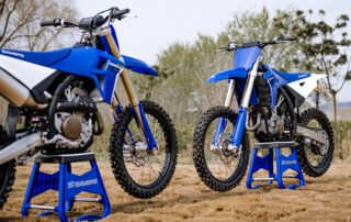 Two blue and white ZXMOTO motocross motorcycles on blue maintenance stands beside a dirt track.