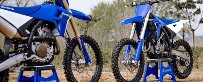 Two blue and white ZXMOTO motocross motorcycles on blue maintenance stands beside a dirt track.
