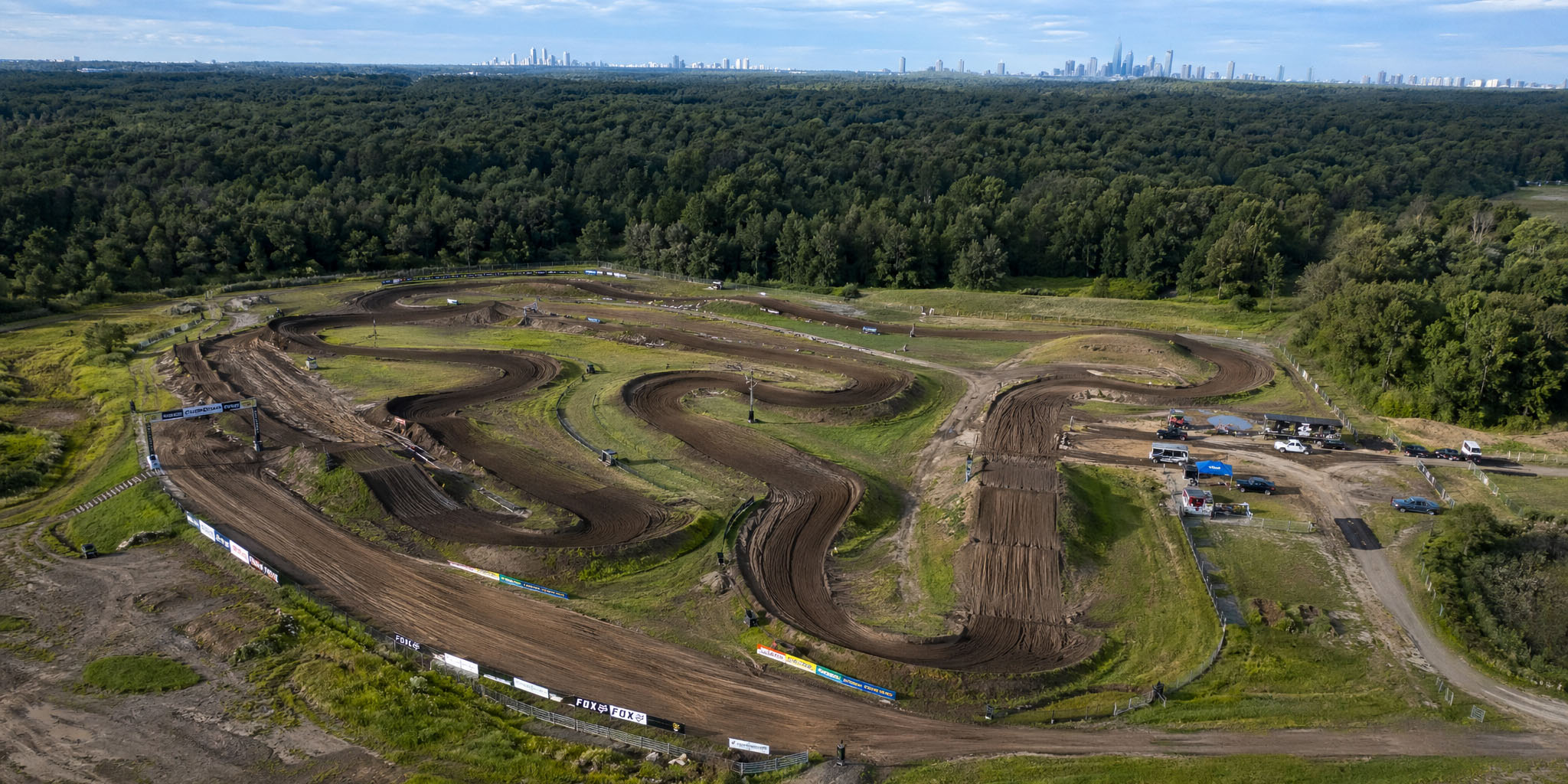 Aerial view of a winding dirt motocross circuit surrounded by woodland, with a distant city skyline.