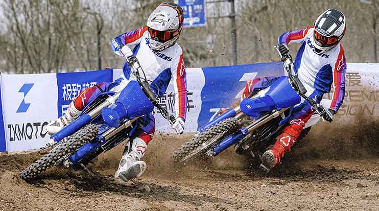Two motocross riders leaning ZX450 dirt bikes through a track corner, kicking up soil beside ZX Moto banners.